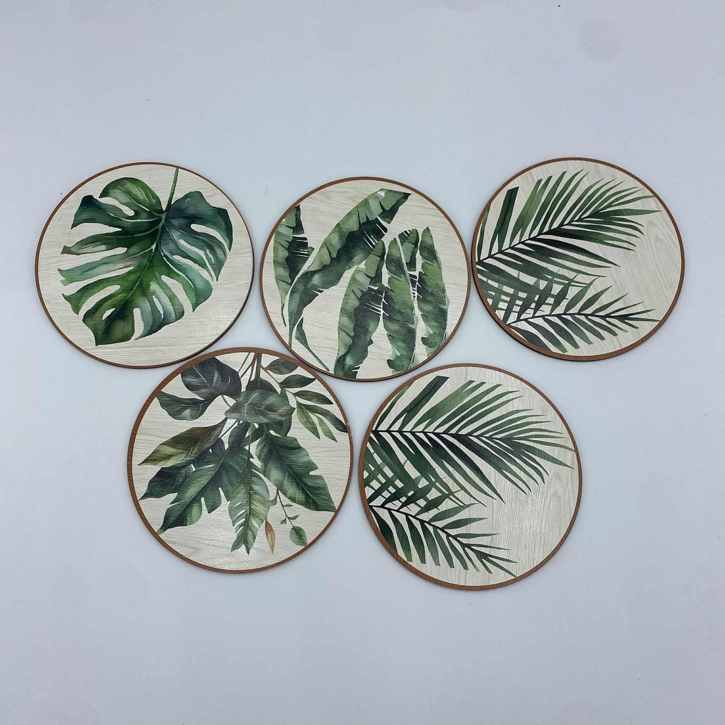  Un ensemble de cinq sous-verres ronds avec des motifs de feuilles de plantes tropicales, dont une feuille de Monstera, sur un fond effet bois clair